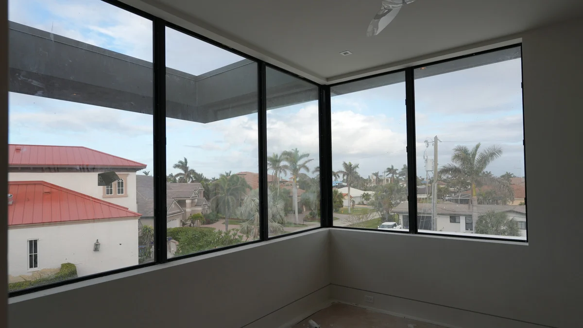 Uncovered corner windows in an unfinished sitting room overlooking a red-roofed neighboring house