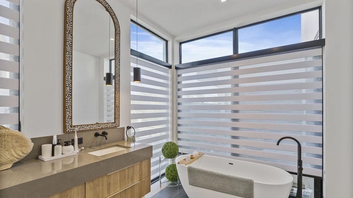 Zebra shades across corner windows behind the freestanding bathtub and finished vanity